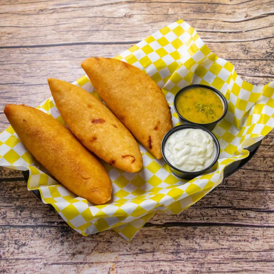 Three golden-fried Venezuelan empanadas in a basket with yellow checkered paper, served with two dipping sauces.