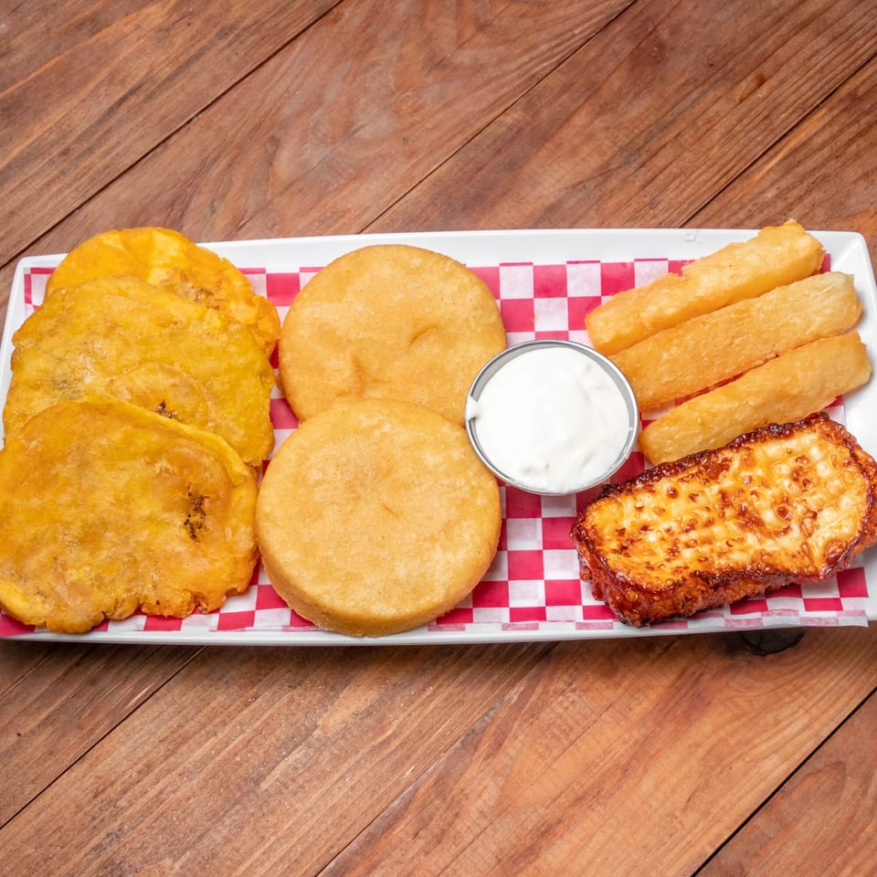 A white platter with Venezuelan food: fried plantains, arepas, yuca fries, grilled cheese, and white dipping sauce.