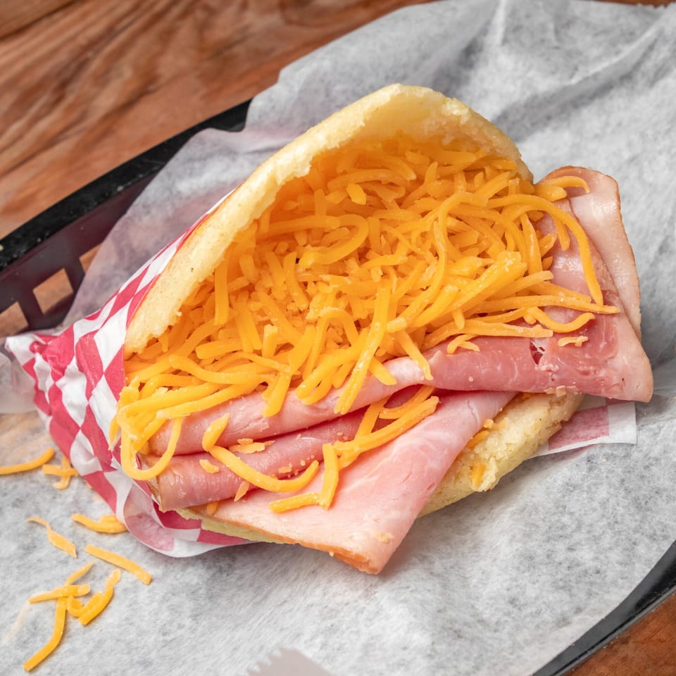 Venezuelan arepa filled with shredded yellow cheese and slices of ham, served in a basket.