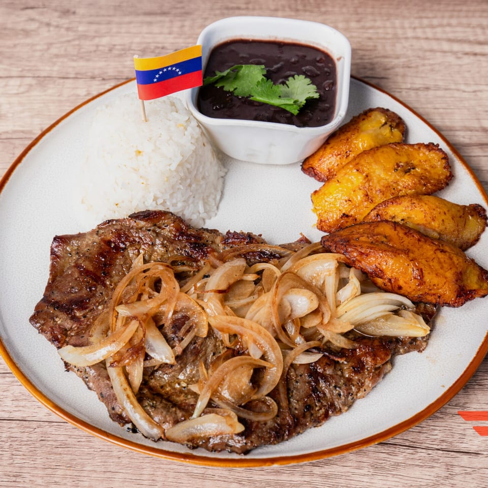 Venezuelan grilled steak with onions, white rice, black beans, fried plantains, and a Venezuelan flag.