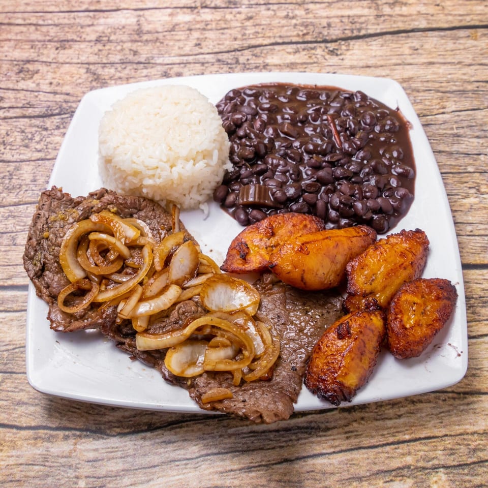 Venezuelan dish with steak and onions, rice, black beans, and fried plantains on a white plate.
