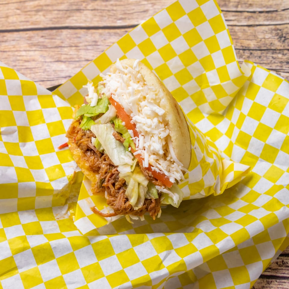 Venezuelan arepa with shredded beef, lettuce, tomato, and cheese, wrapped in yellow checkered paper.