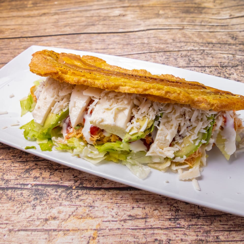 A Venezuelan patacon with shredded chicken, avocado, cheese, and lettuce on a white plate.