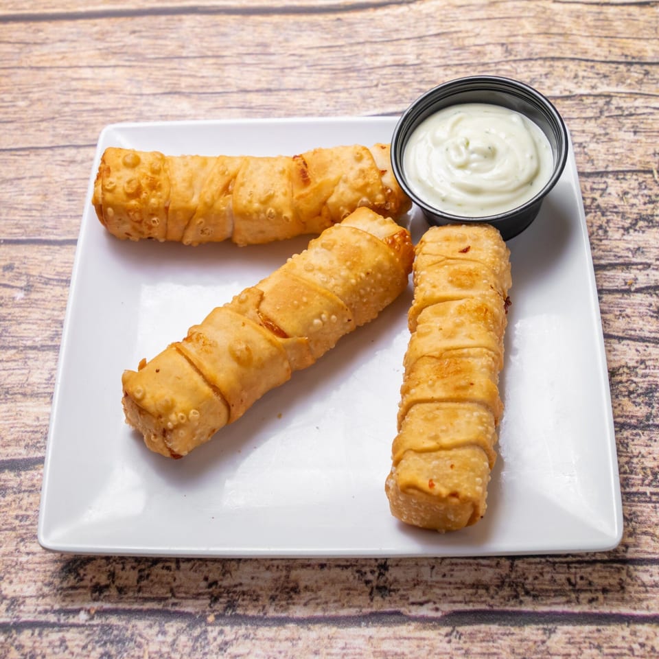 Three golden-brown Venezuelan tequeños, fried cheese sticks, on a white plate with a side of creamy white sauce.