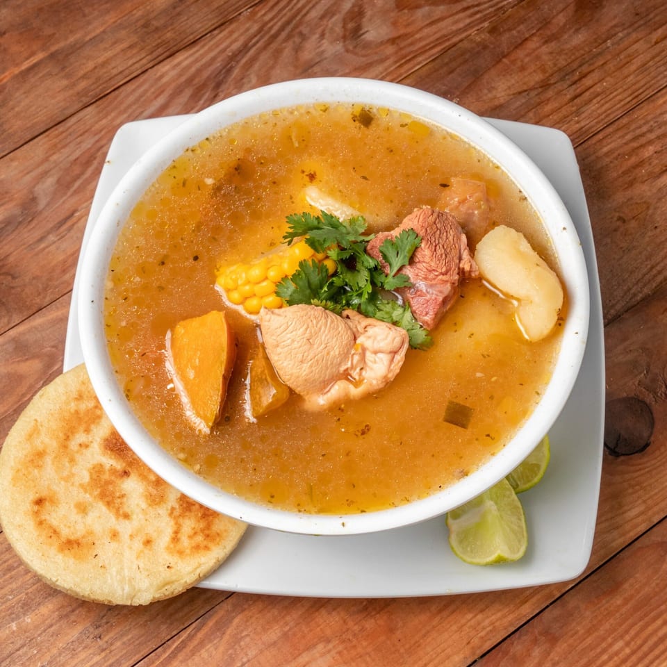 Venezuelan beef soup with corn, potatoes, and cilantro, served with an arepa and lime wedges.