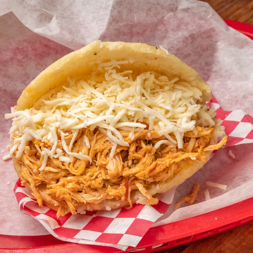 Close-up of a Venezuelan arepa filled with shredded chicken and white cheese, served in a red basket.