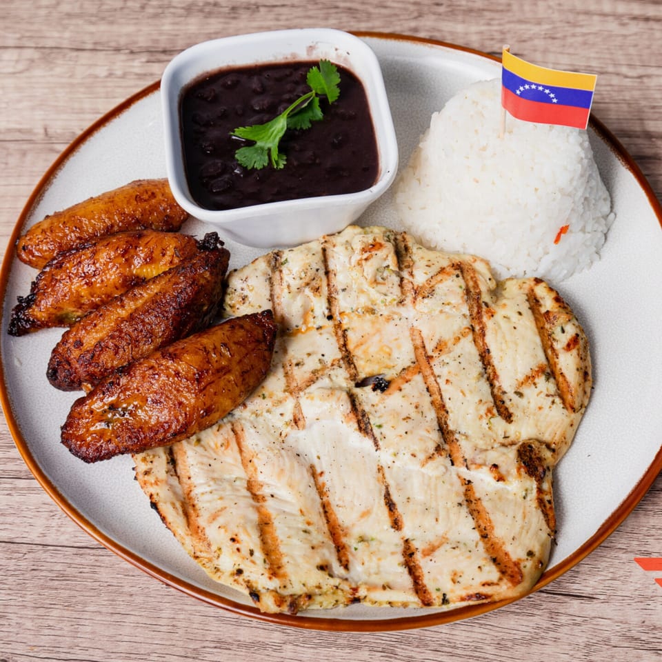 Venezuelan grilled chicken, black beans, rice with a flag, and fried plantains on a plate.