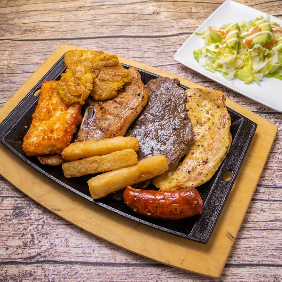 Venezuelan mixed grill with fried pork, steak, chicken, sausage, yuca fries, and a side salad.