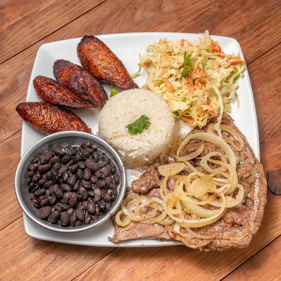Venezuelan dish with steak, onions, rice, black beans, fried plantains, and coleslaw on a white plate.